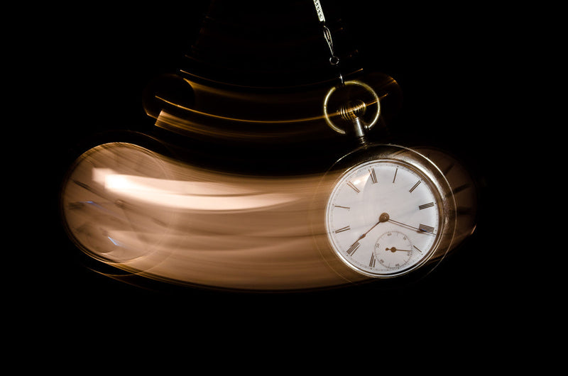 A blurred pocket watch swings in motion against a dark background, evoking the pull of time and unseen forces. Part of Candle Face Chronicles.