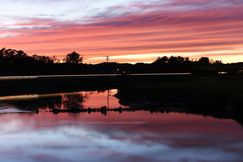 A sunset casts vibrant pink and orange hues across the sky, reflecting on the calm waters below. A road in the distance is lined with trees, and streaks of light from passing vehicles hint at motion. The image conveys a sense of mystery and contemplation,