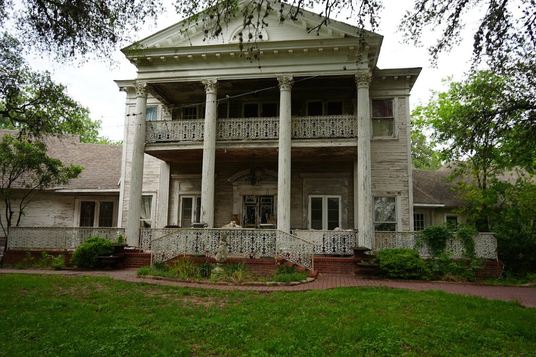 An old, decaying mansion with white pillars and wrought-iron railings, surrounded by overgrown greenery. Represents 'Preparing for Candle Face’s Return,' from Candle Face Chronicles by Branching Plot Books.