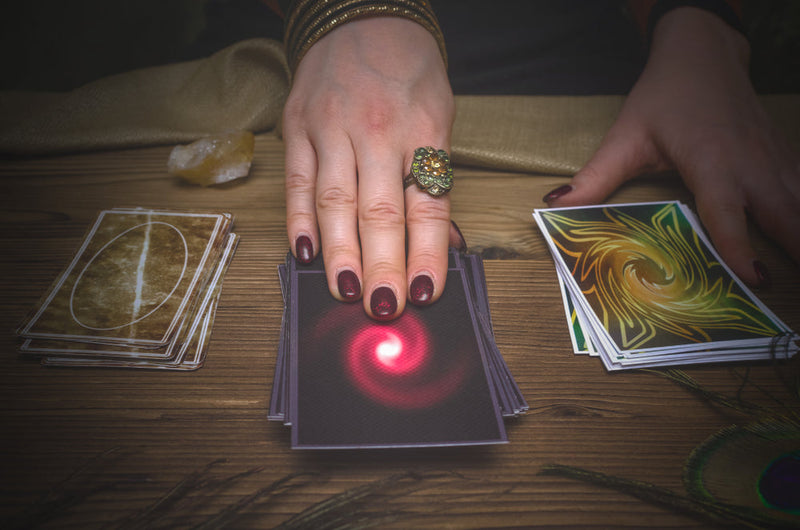 The image shows a psychic's hand with elegantly painted nails and a decorative ring, placing or drawing a tarot card with a glowing red spiral design from a deck laid out on a wooden table. Other cards with swirling designs are visible.