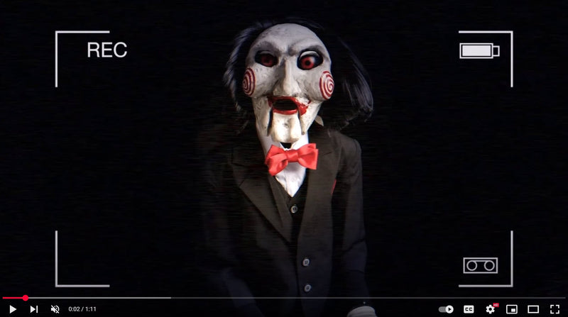 Video screenshot of a puppet-like figure with a white mask, red spiral cheeks, and black hair, wearing a black suit with a red bowtie. Image titled 'The Empty Lot Next Door: Arthur Mills' Haunting Childhood.