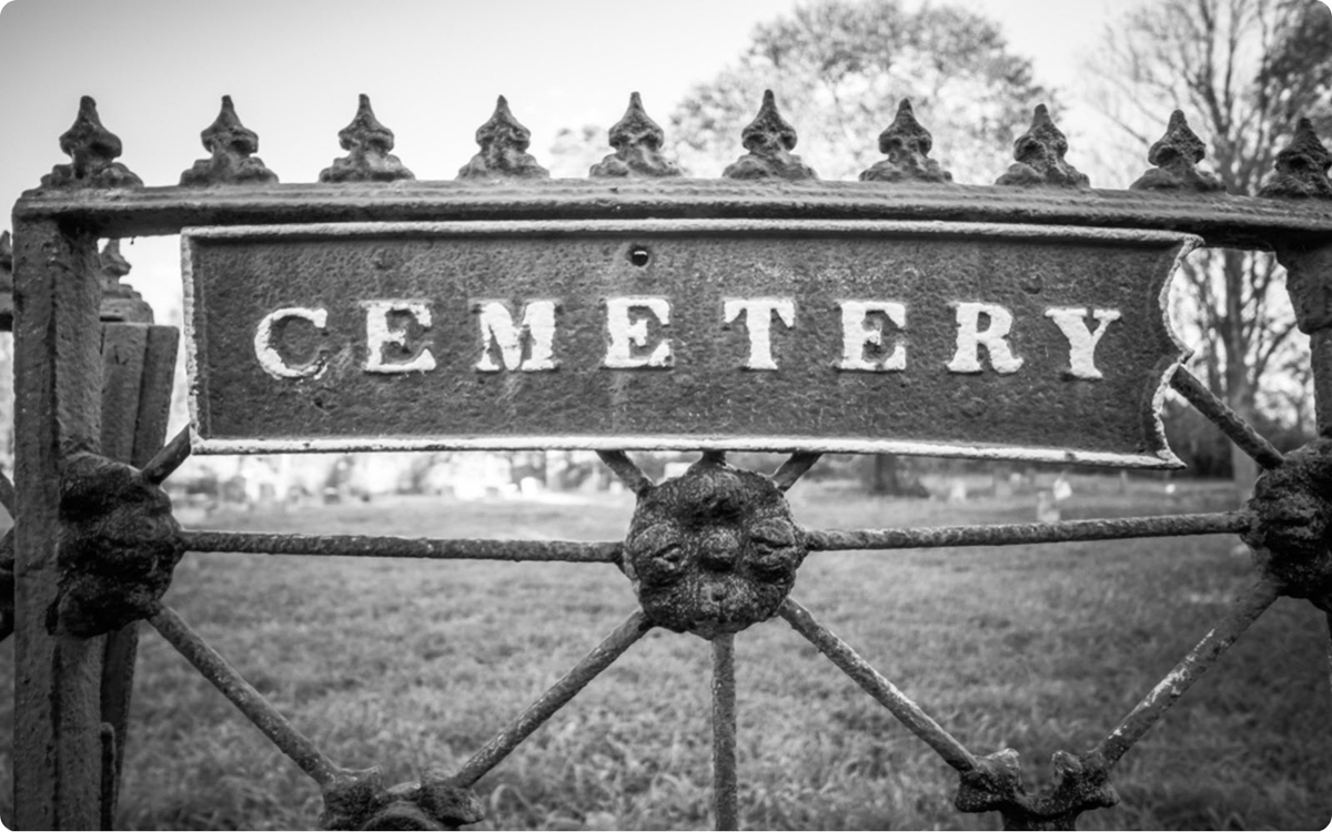 A rusted iron gate with the word 'CEMETERY' in bold, weathered lettering—an entryway to forgotten graves and lost stories waiting to be rediscovered with Branching Plot Books.