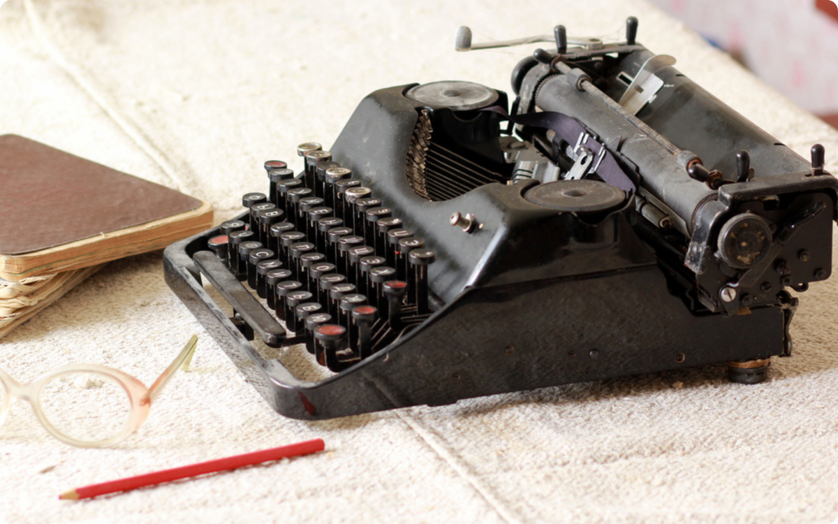 A vintage black typewriter sits on a textured surface beside a worn notebook, a red pencil, and reading glasses. Symbolizing storytelling and the craft of writing, this image represents the dedication behind Branching Plot Books and its interactive nature.