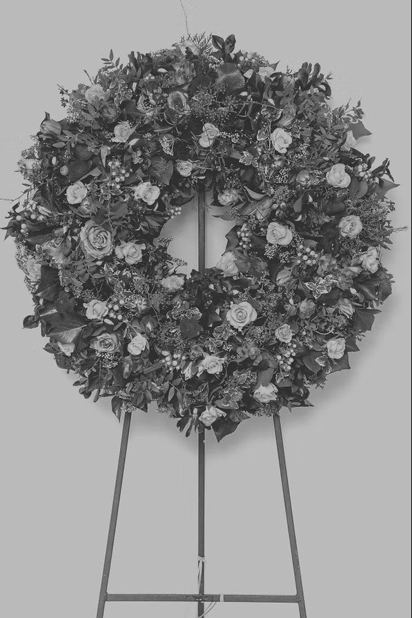 A somber, monochromatic floral wreath displayed on a metal stand. The wreath is composed of roses, leaves, and berries, arranged intricately to create a hauntingly beautiful yet melancholic display. This image is associated with Candle Face Chronicles and