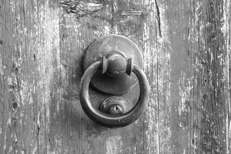 A weathered, rustic door with a round, metal knocker against a worn wooden surface. The image symbolizes the tragic story of 'Candle Face Victim 45: The Friend Who Didn't Answer,' part of the Candle Face Chronicles by Branching Plot Books.