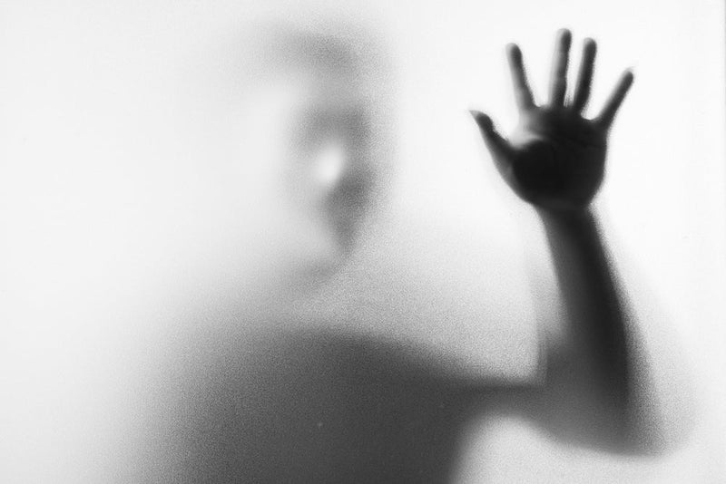 A blurred, shadowy figure presses a hand against a frosted glass or fogged surface, the details obscured but the gesture seemingly desperate or haunting. This image represents the elusive nature of the Fugitives trying to reach out for help while still re