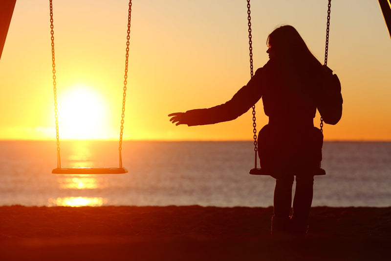 Silhouette of a woman on a swing at the beach during sunset, reaching toward an empty swing beside her. Image titled 'IDENTIFIED? Candle Face Victim #13: The Drowning Ghost' from Candle Face Chronicles.