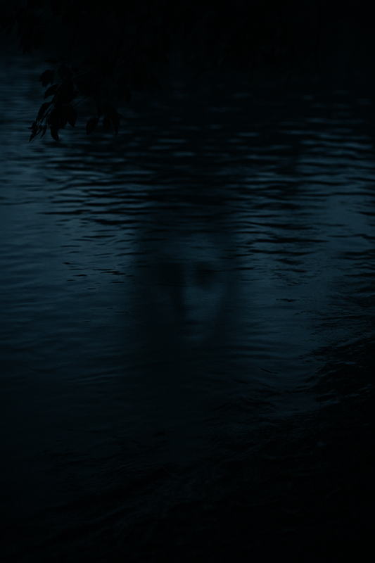 A dark, moody view of Lady Bird Lake at night. The water’s surface is still, with faint ripples. Reflected in the water is a barely visible, ghostly face or shadow, its features soft and indistinct, evoking the eerie presence of Isabel.
