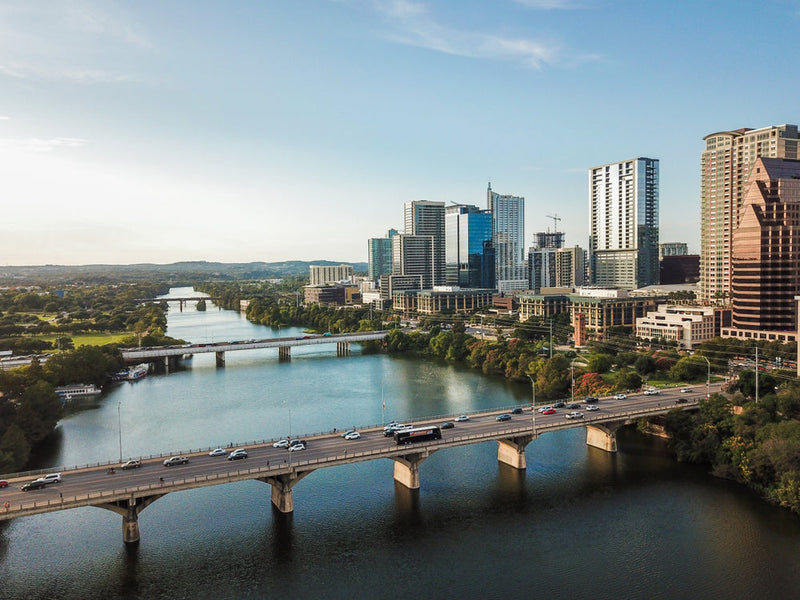 An aerial view of Congress Avenue Bridge in Austin, Texas, with modern skyscrapers rising against a clear blue sky. The calm waters of the river below reflect the surrounding structures and traffic moving along the bridge. This image, titled 'Pushed from 