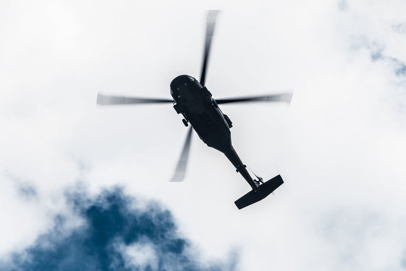 A dark helicopter flies overhead against a cloudy sky, its rotor blades spinning. The ominous angle and shadowed silhouette suggest secrecy and surveillance. Related to Candle Face Chronicles.