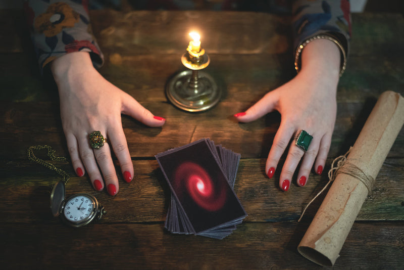 A dimly lit wooden table holds a deck of tarot cards, a candle in a brass holder, an old pocket watch, and a rolled parchment tied with twine. A woman with red-painted nails and ornate rings on both hands rests them on the table, poised as if ready to con
