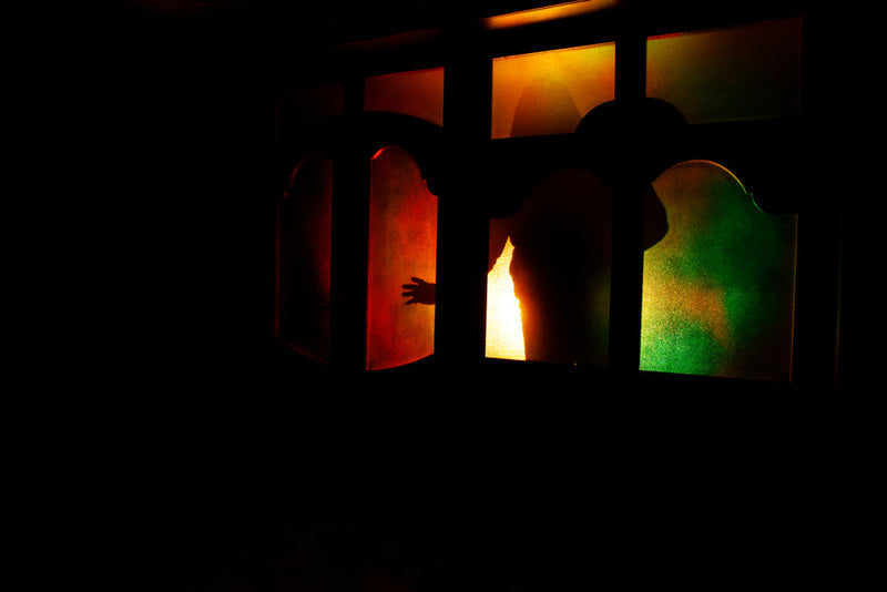 A shadowy figure reaching through colorful, stained glass panels illuminated by glow of red, yellow, and green. The image symbolizes the unpredictable and omnipresent nature of the Master Shadow from Candle Face Chronicles by Branching Plot Books.