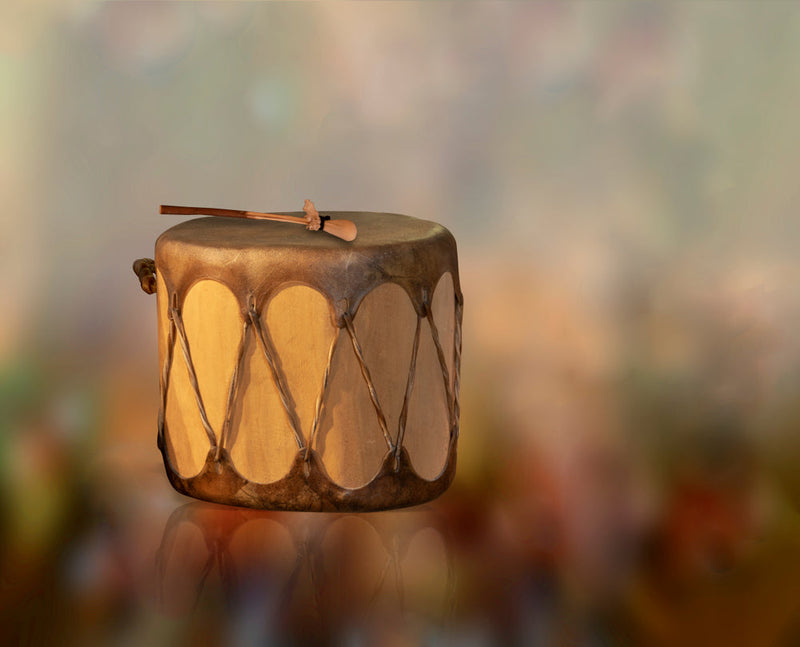 A handcrafted drum with a natural hide surface and intricate leather bindings, resting on a reflective surface against a softly blurred background of earthy tones. The drumstick lies atop the drum, hinting at a ritualistic or spiritual purpose. This image