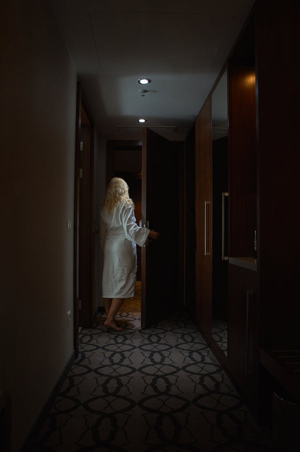 A woman in a white robe walks barefoot down a dimly lit hallway, opening a door into darkness. Image titled 'The Torment of Betrayal.'