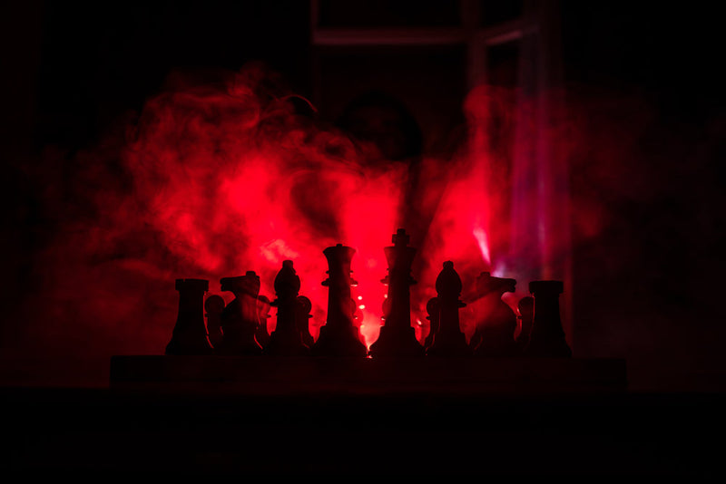 A chessboard sits in red mist, its pieces silhouetted against the glow. In the background, a hooded figure watches from the shadows. This image represents the unsettling power struggle from 'Tour of Torment,' symbolizing strategy, manipulation, and the un