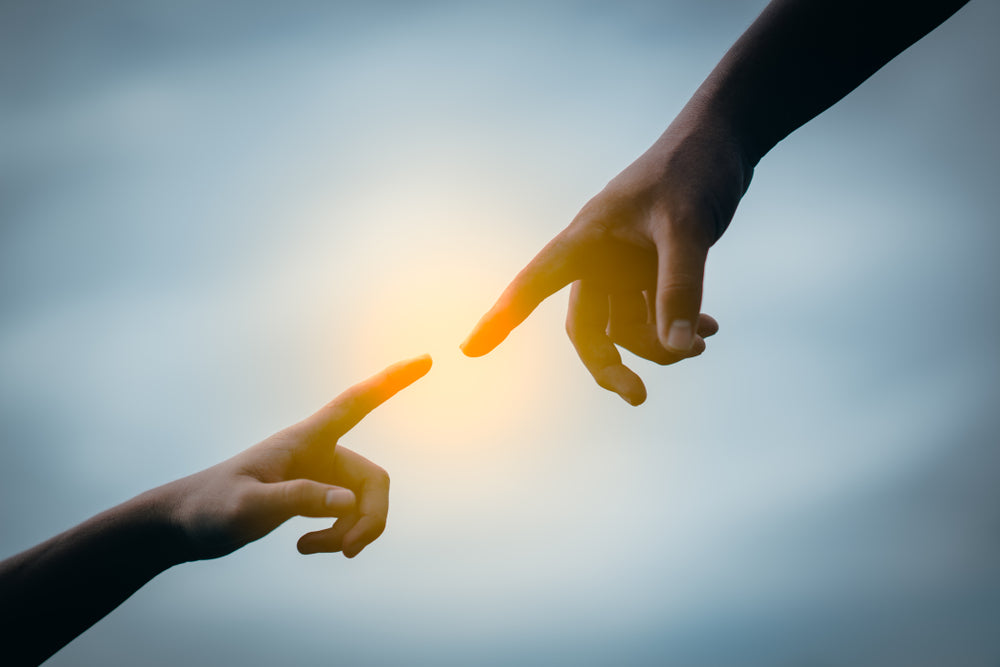 Two hands reaching toward each other with fingers nearly touching, illuminated by a soft glowing light in the center. The image symbolizes the connection between a lost soul and its caretaker in the Scrolls of Souls project.