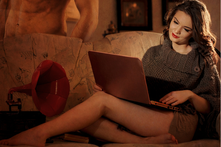 Co-Author by Arthur Mills. Chelsie sits on a sofa typing on a laptop, her shirtless hero imagined standing beside her.