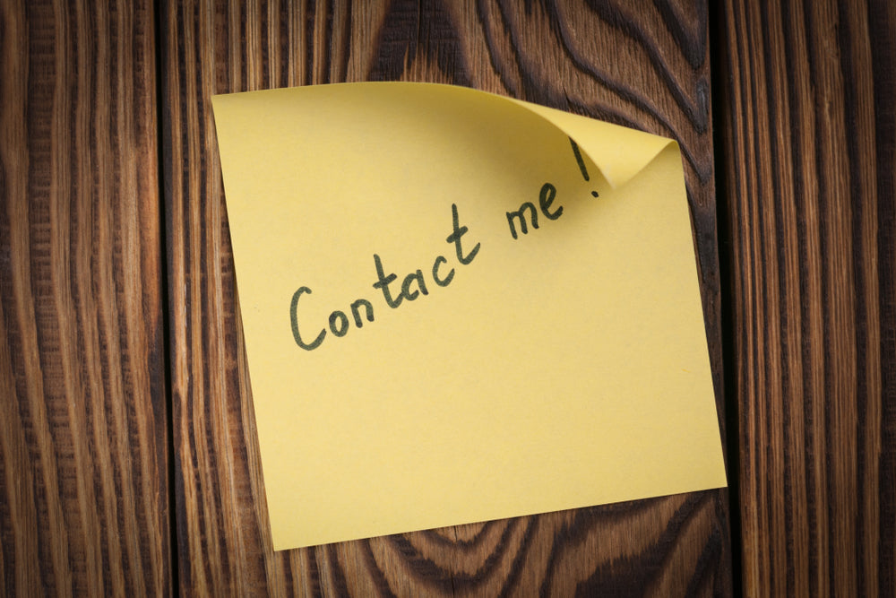 Yellow sticky note with "Contact me!" handwritten, stuck to a wood wall.
