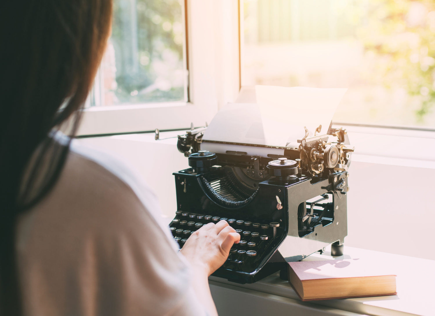 Woman typing on a vintage black typewriter by a sunlit window, a book resting on the desk.